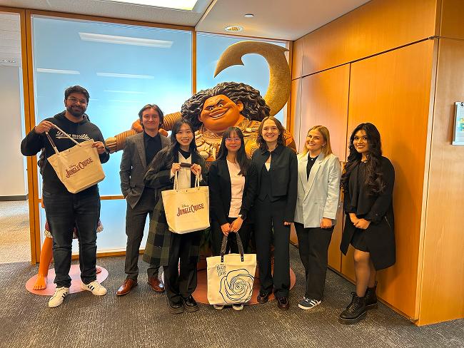 A group of elated students pose in front of a Maui statue from the hit Disney movie Moana holding tote bags